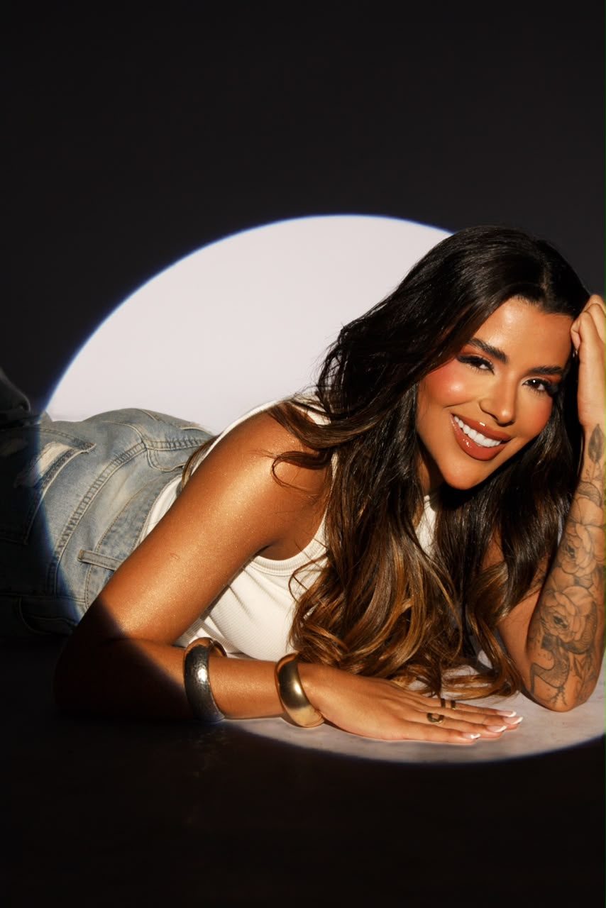 Juliana Cardoso deitada sob um círculo de luz, de regata branca e jeans, sorrindo para a câmera